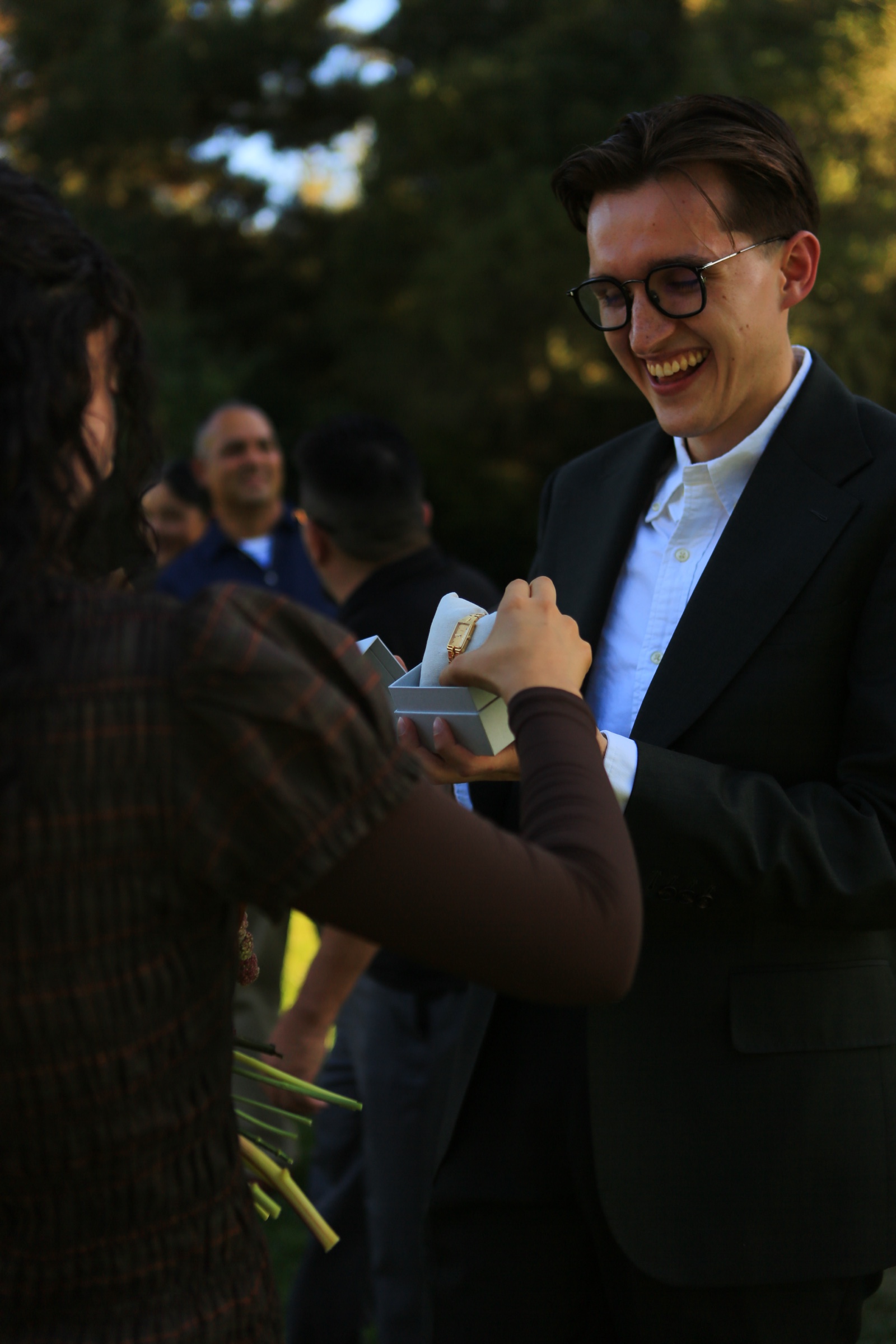 A man in glasses smiles while holding an open watch box as a woman reaches toward the gold-colored watch inside.