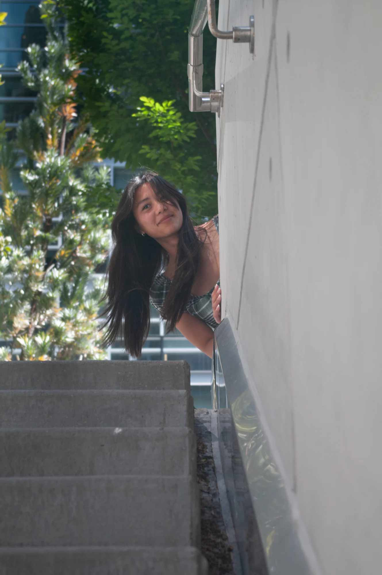 Woman peeking around a white wall beside outdoor steps and greenery.