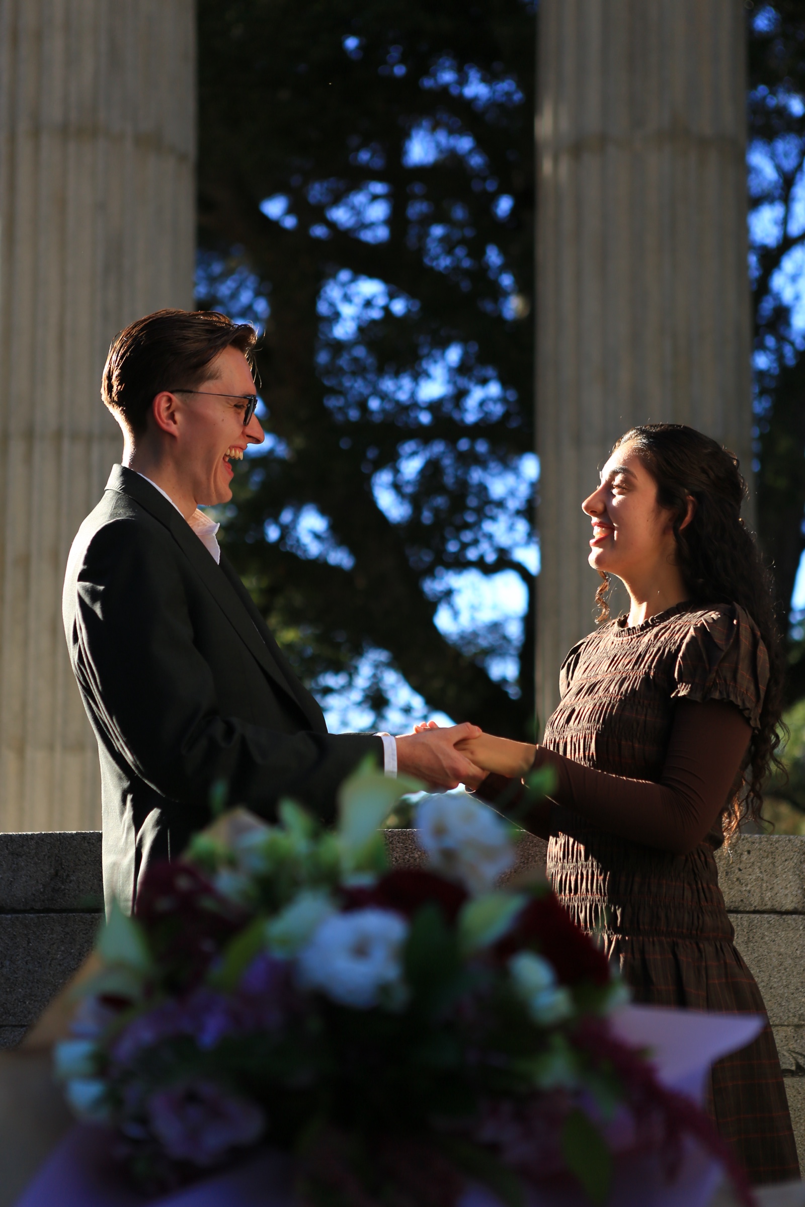 A smiling couple holds hands between stone columns, with a softly blurred bouquet in the foreground.