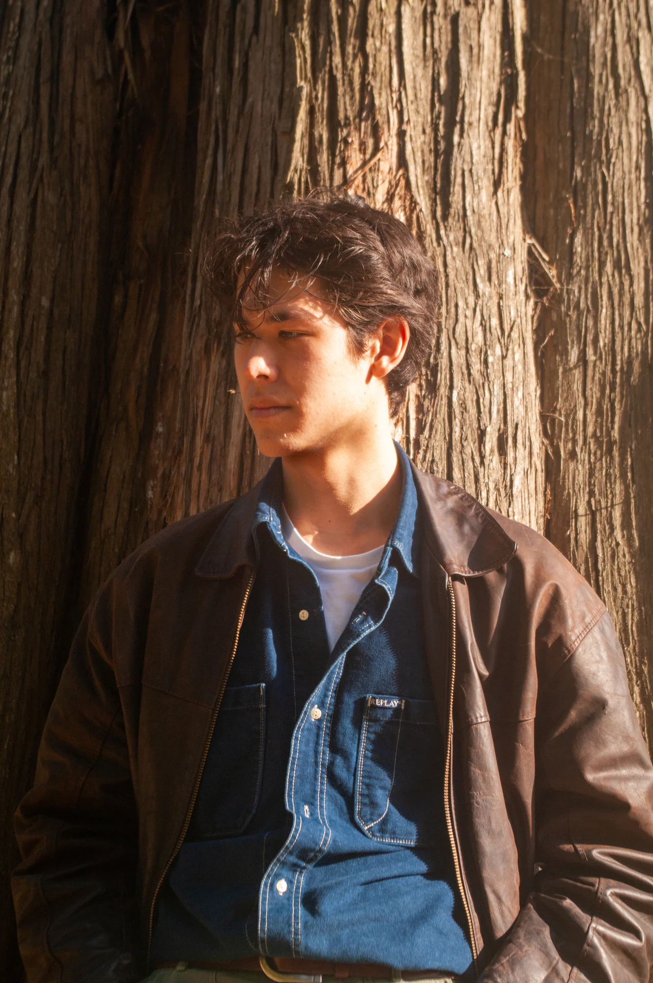 Young man in layered denim and leather standing against a sunlit tree.