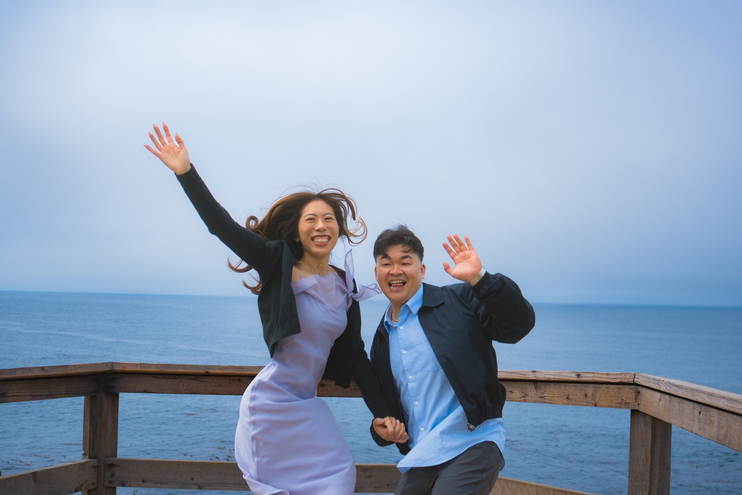 Newly engaged couple celebrating with raised hands at an ocean overlook.
