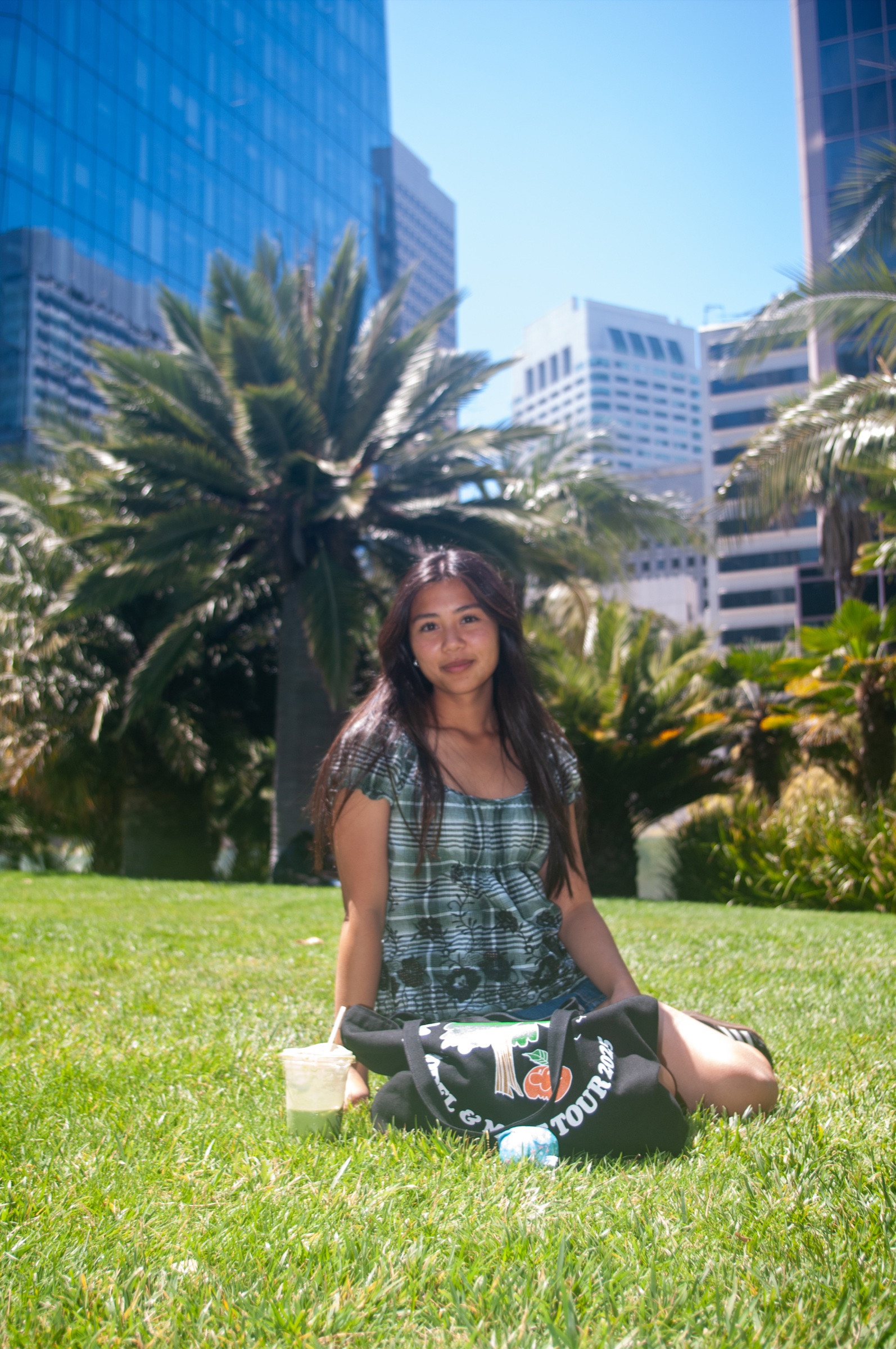 Sunlit portrait of a woman seated on green grass among palms and downtown buildings.