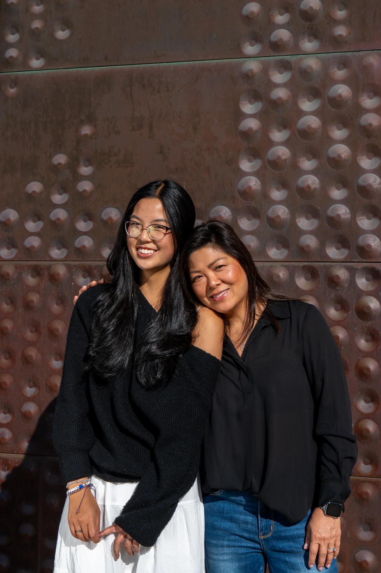 Two women smiling together in bright sunlight against the de Young Museum's textured copper wall.