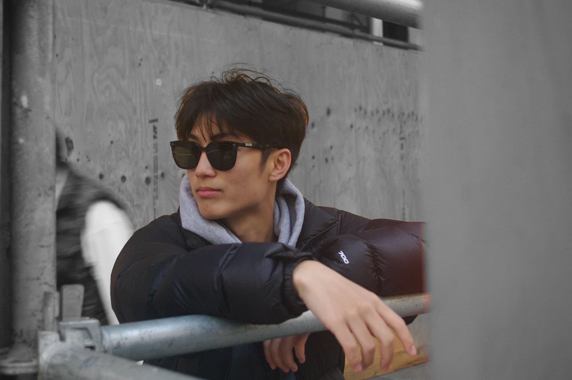 Man in dark sunglasses and a black jacket leaning against a rail in front of a concrete wall.