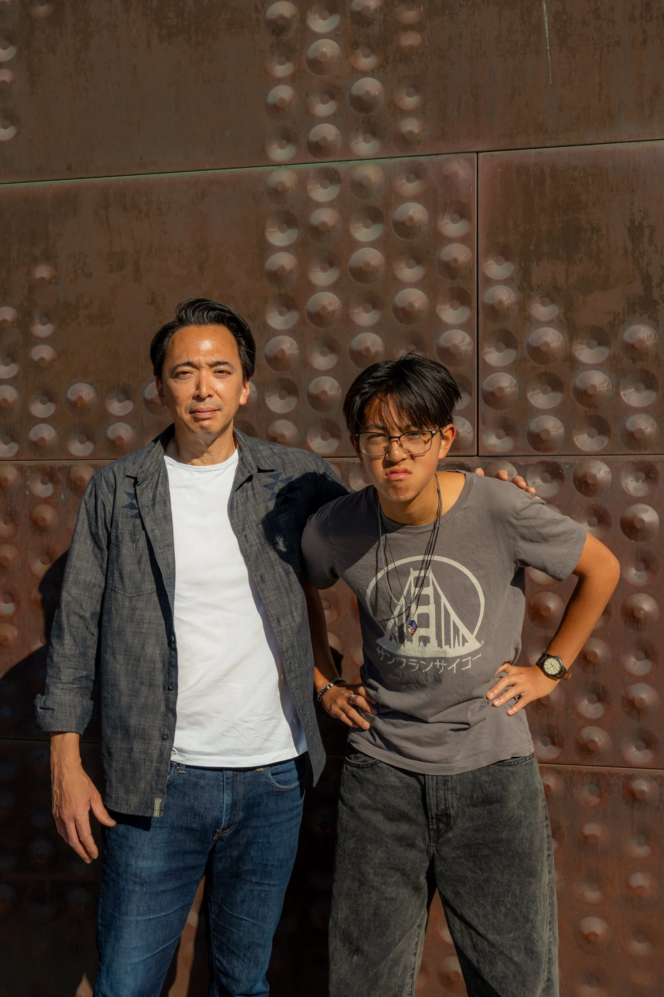 Two men posing together in bright sun against the de Young Museum's textured copper wall.