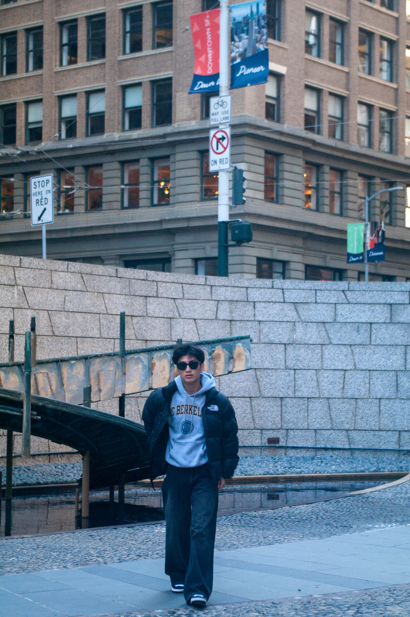 Young man in sunglasses and a Berkeley hoodie walking through a downtown plaza.