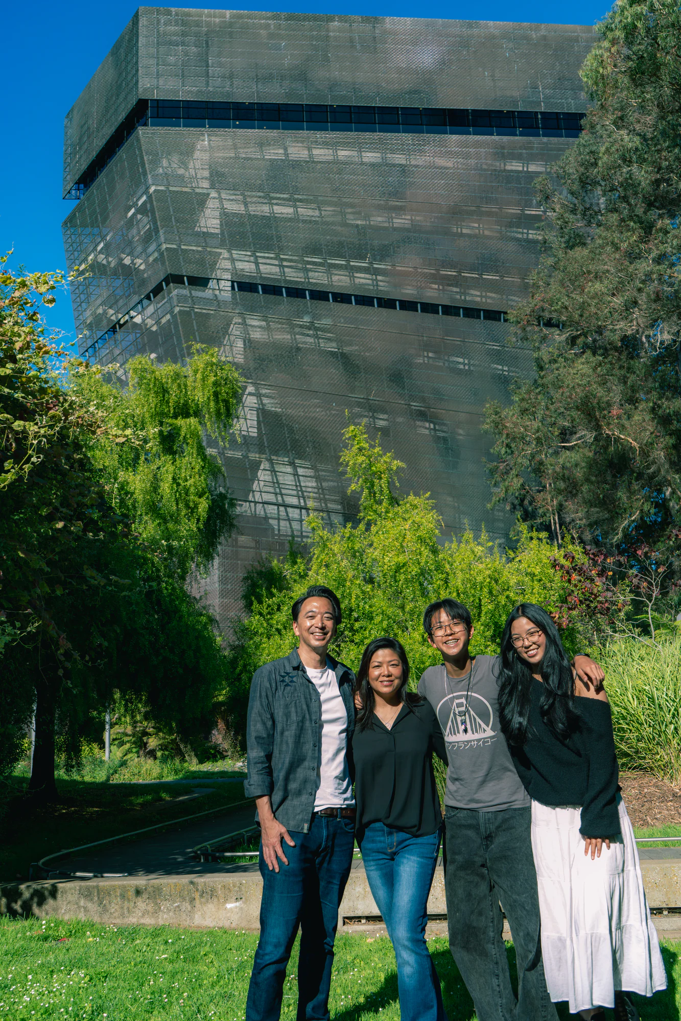 Family of four smiling outdoors beneath the de Young Museum tower and a vivid blue sky.