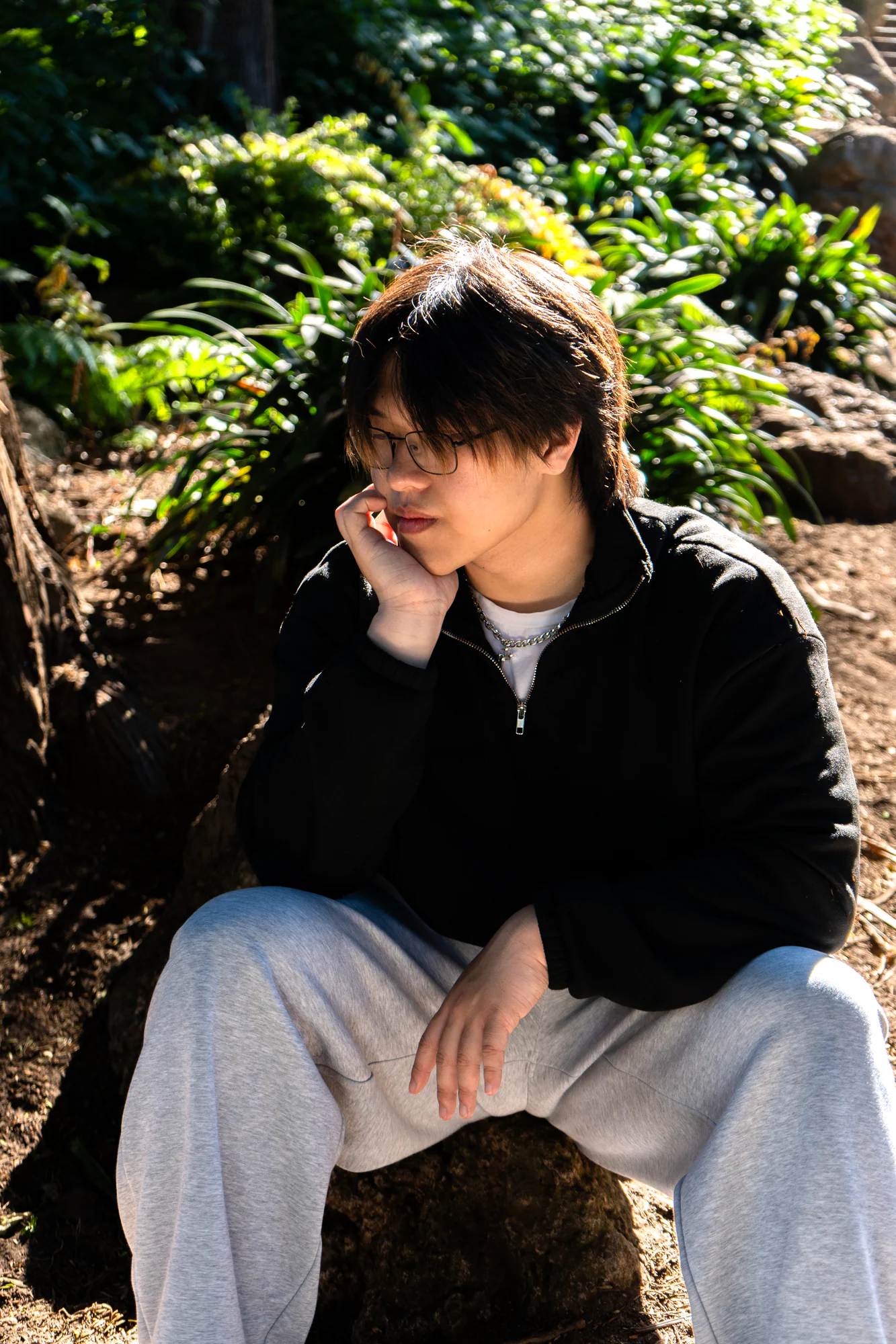 Portrait of a person in glasses sitting among sunlit trees with their chin resting on one hand.