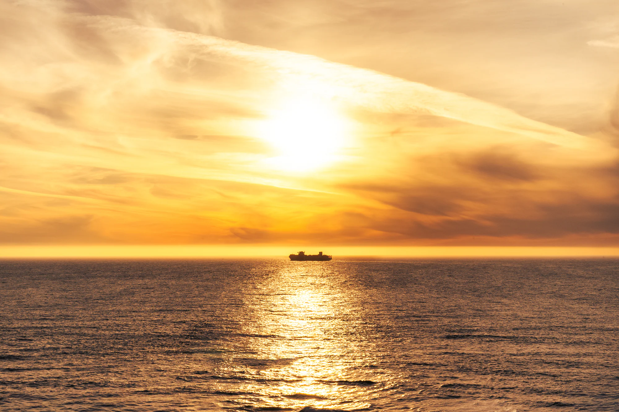Cargo ship silhouetted beneath a glowing gold sky, with sunlight reflecting across the ocean.