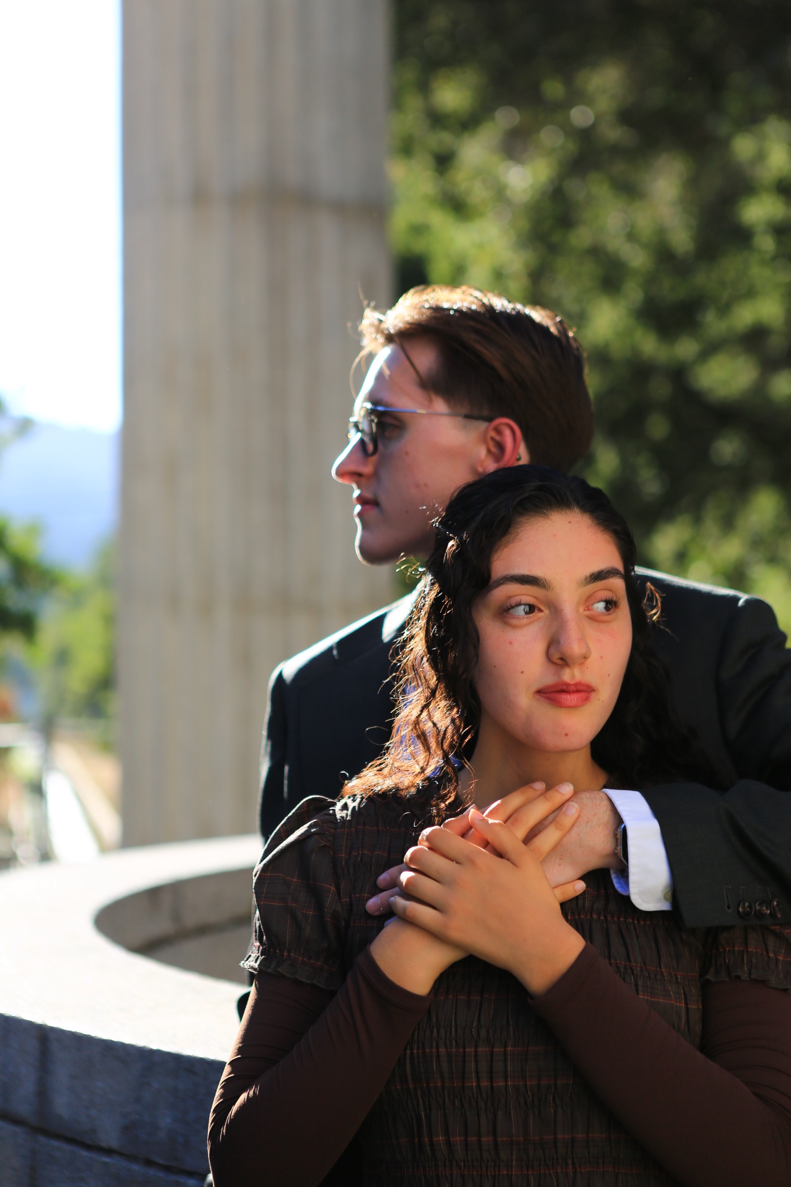 A woman rests her hands over her partner's arm as he stands close behind her beside a sunlit stone column.