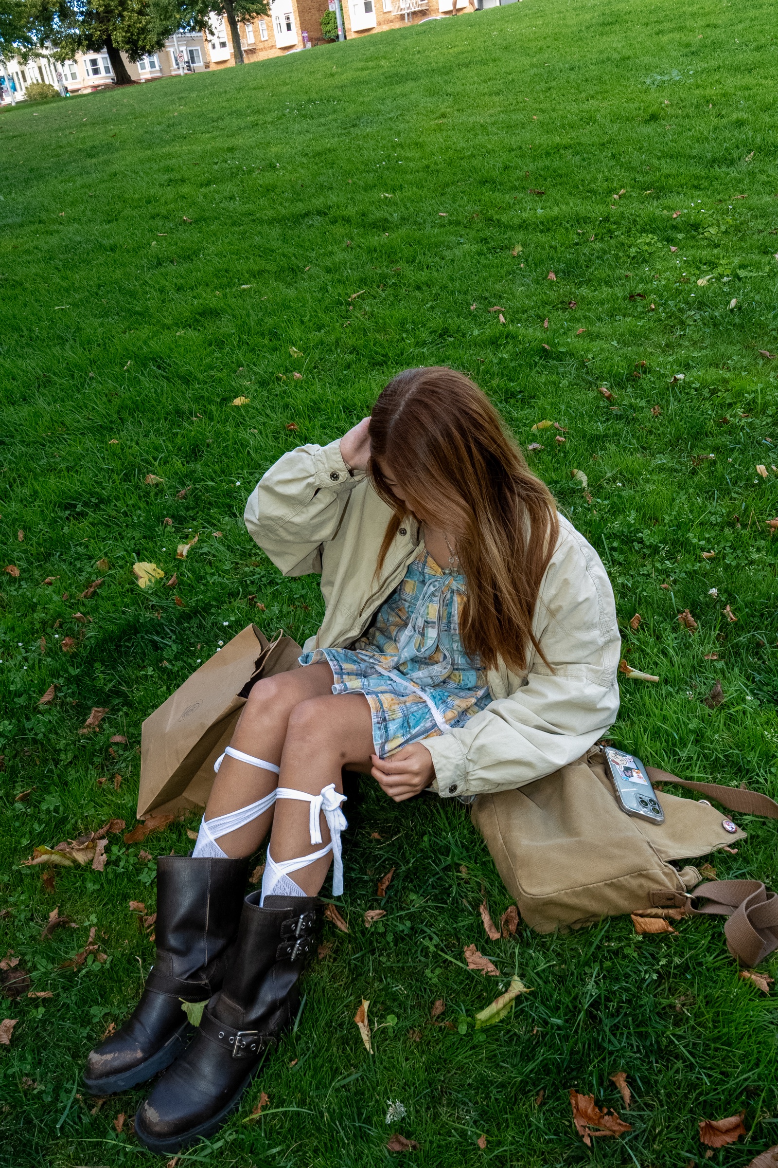 Candid portrait of a woman seated on bright green grass in a plaid dress and oversized jacket.