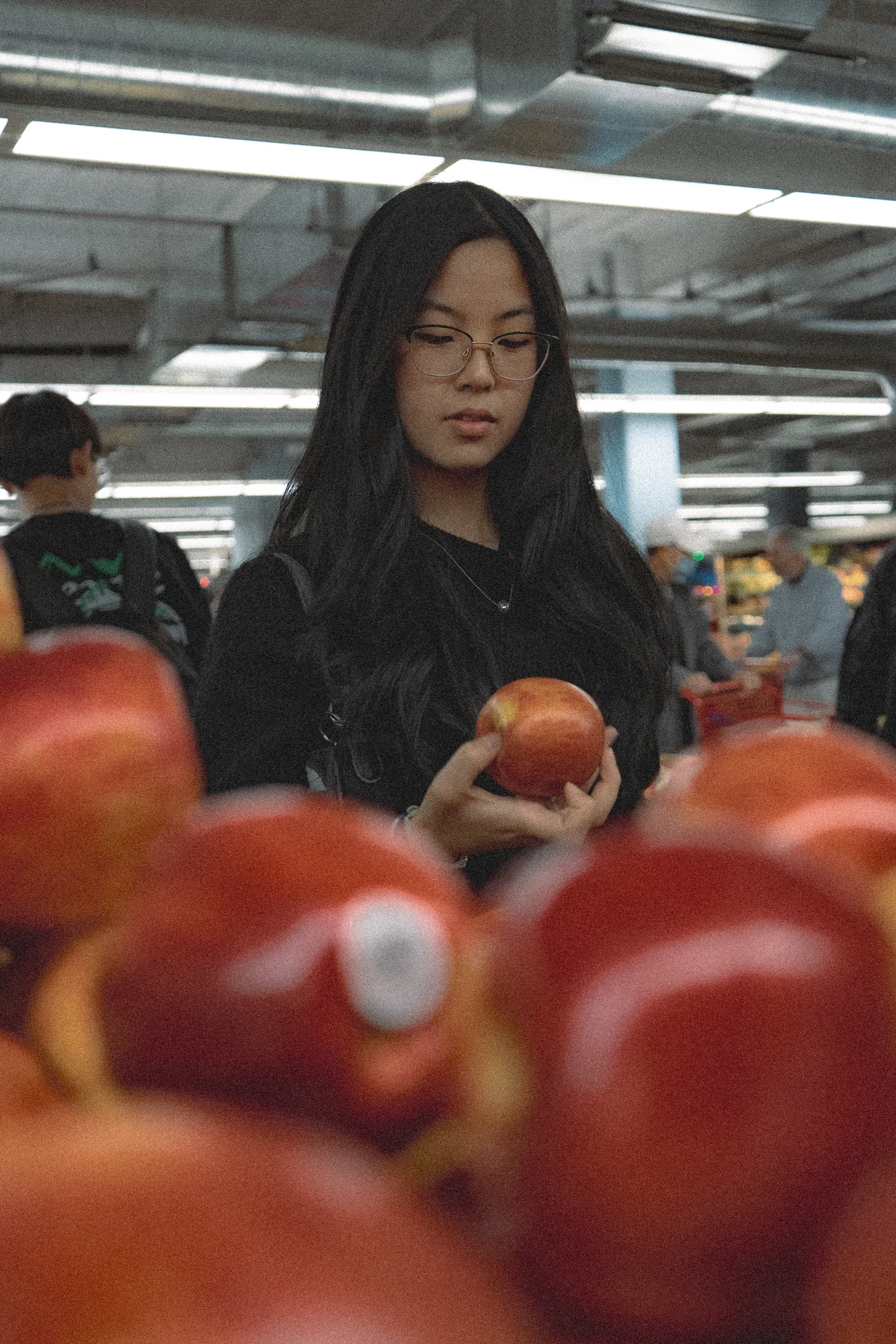 Woman with glasses holding a red apple among produce in a brightly lit market.