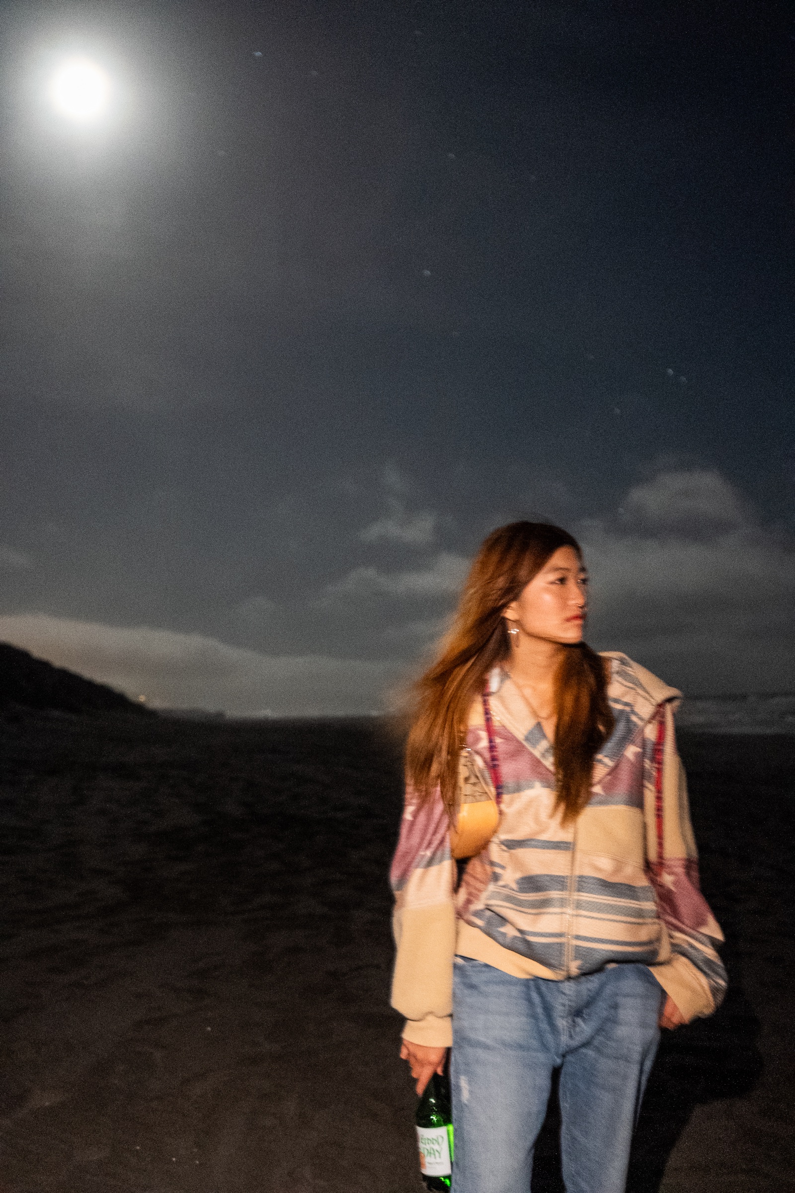 Night portrait of a woman beneath a bright moon wearing a colorful jacket and holding a bottle.
