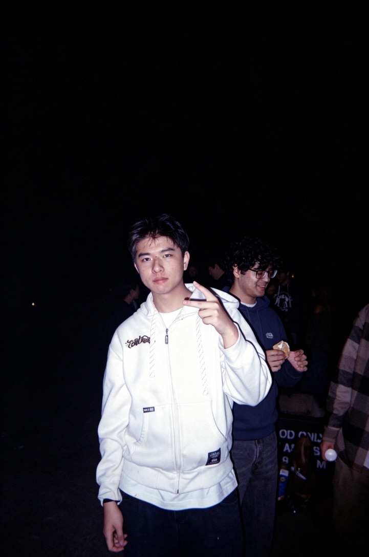 Flash portrait of a young man in a white hoodie making a peace sign at an outdoor gathering after dark.