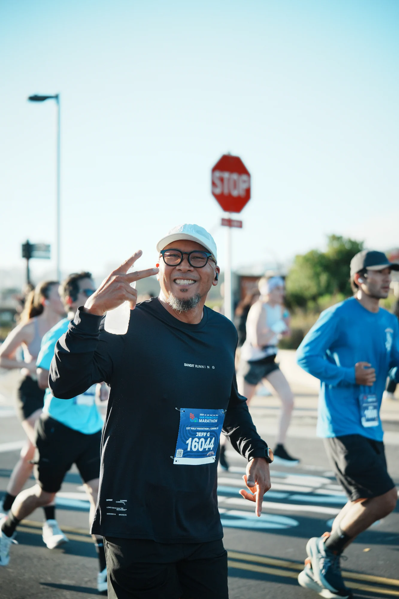 Runner in a white cap and black shirt smiles at the camera and flashes a peace sign during the San Francisco Marathon.