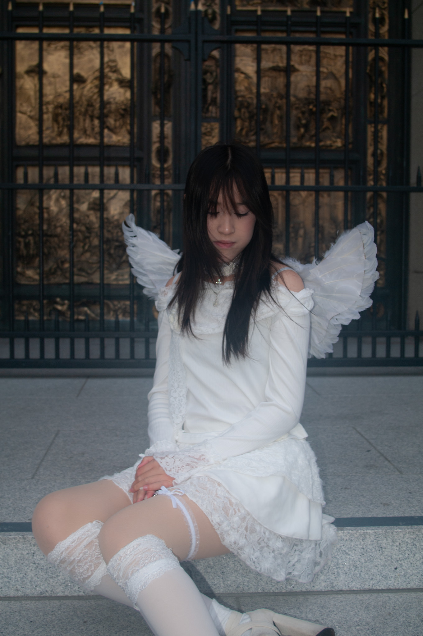 Editorial portrait of a woman in a white angel costume seated on stone steps in front of ornate metal doors.