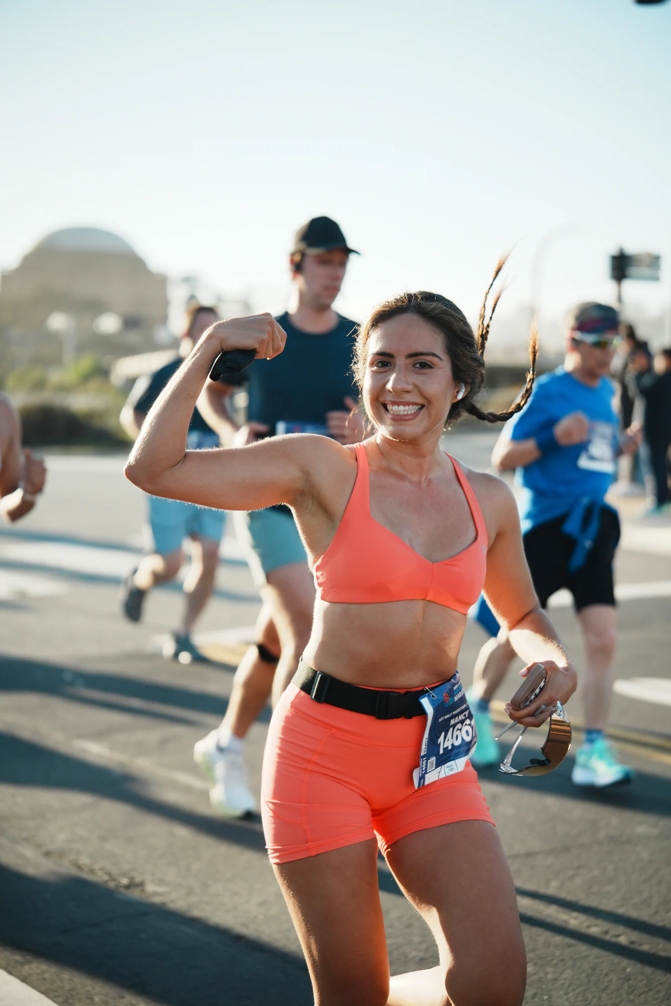 Smiling runner in coral racewear flexes one arm during the San Francisco Marathon as other runners pass behind her.