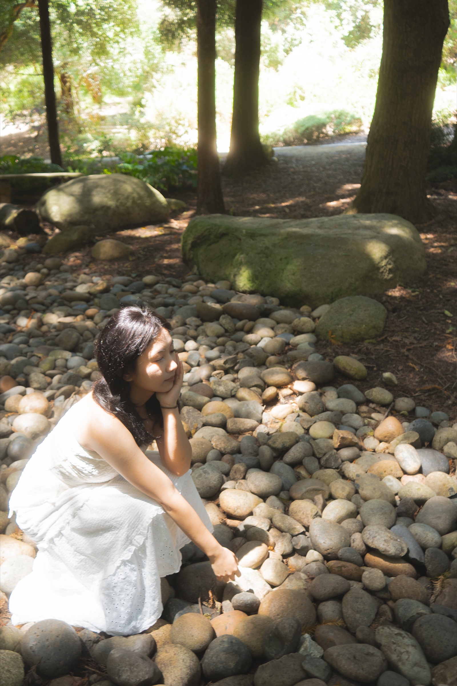 Portrait of a woman in a white dress seated among sunlit stones beneath trees in a shaded garden.
