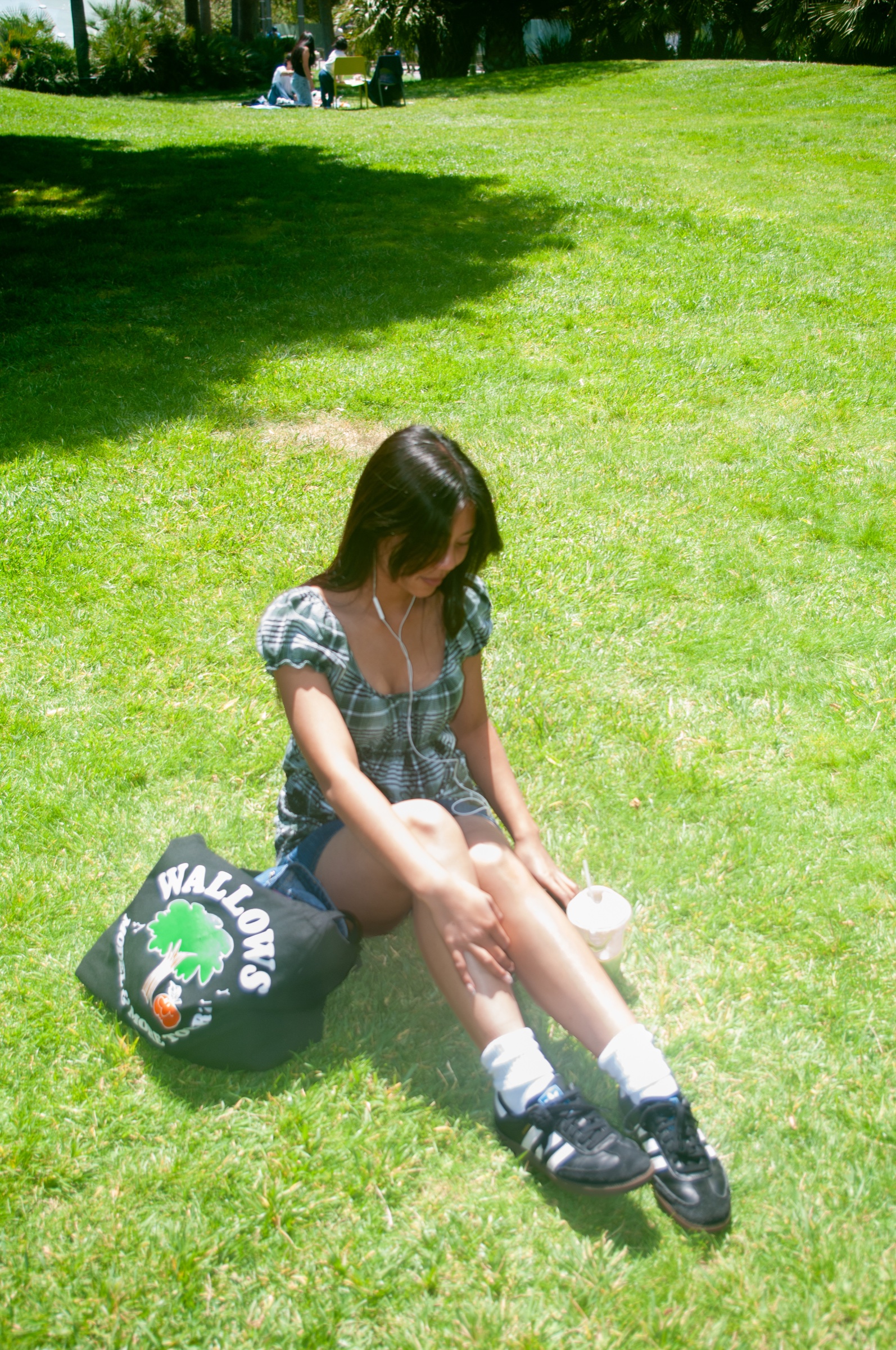 Candid sunlit portrait of a woman seated on a bright green lawn with headphones and a tote bag.