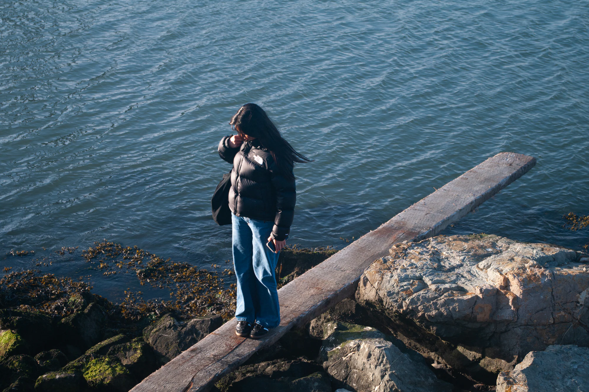 Person in a black puffer jacket standing on rocks beside blue water.
