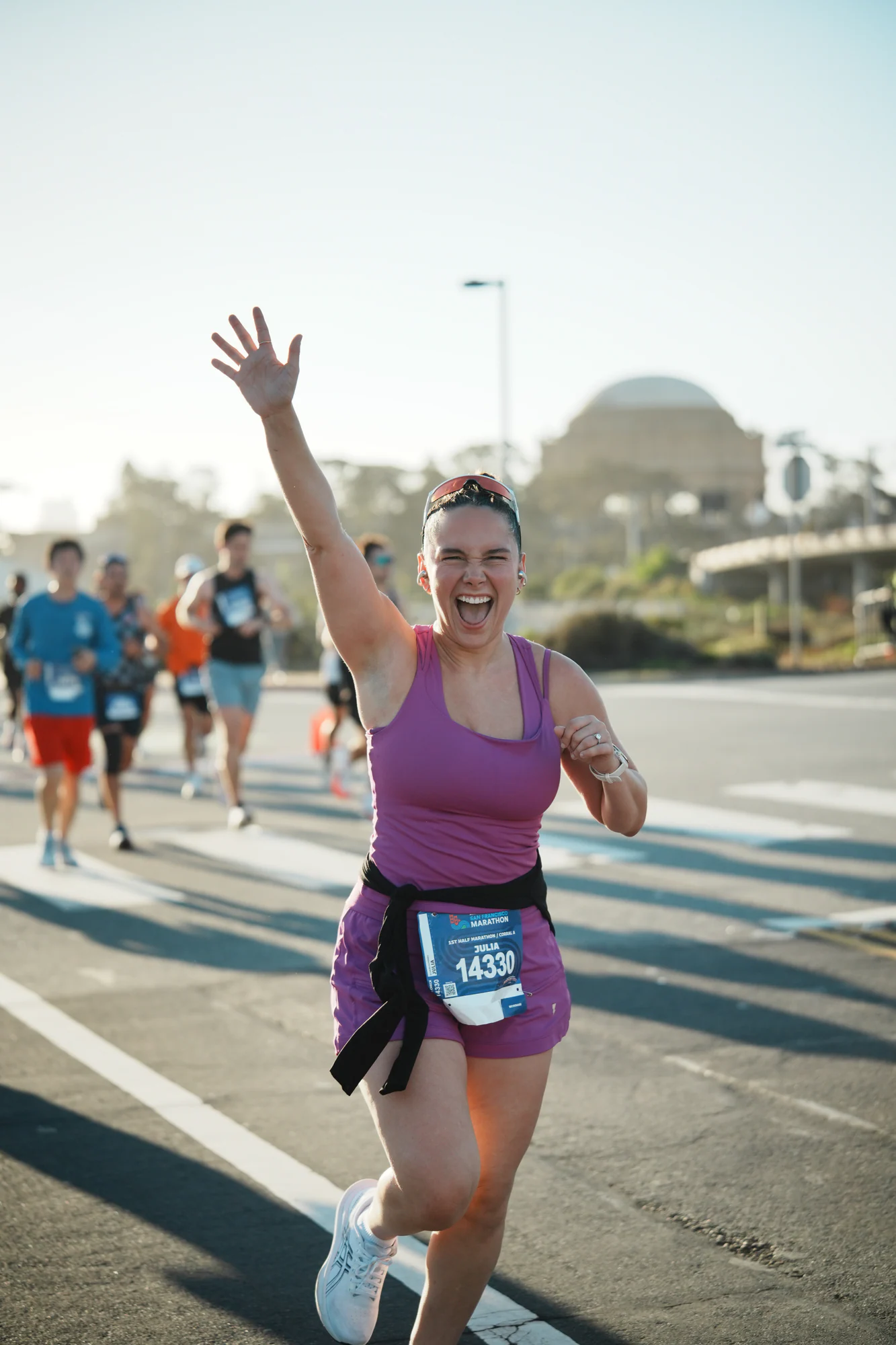 Runner in purple racewear waves overhead and smiles broadly on a sunlit San Francisco Marathon course.